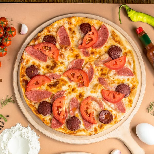 A Mediterranean pizza with salami, sausage, and tomato slices on a wooden board, surrounded by ingredients.