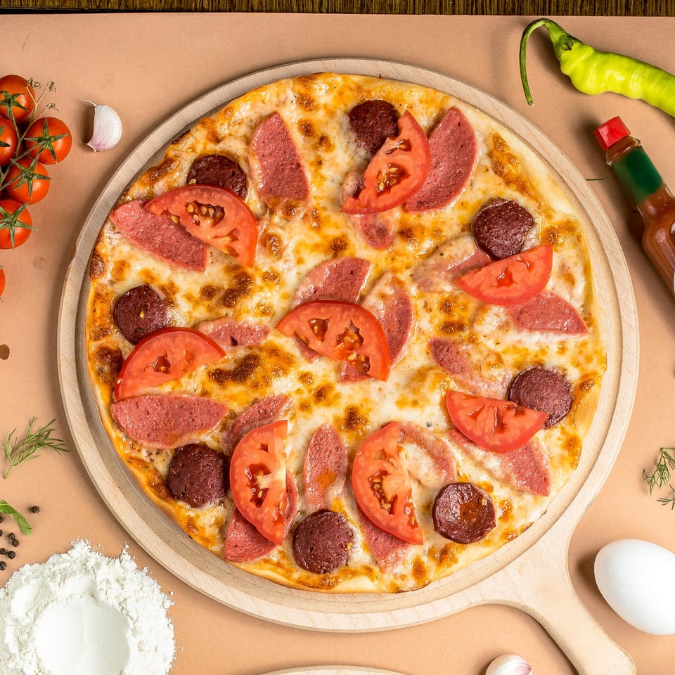 A Mediterranean pizza with salami, sausage, and tomato slices on a wooden board, surrounded by ingredients.