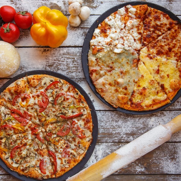Two pizzas, one with vegetables and one with four cheeses, on a flour-dusted wooden table.
